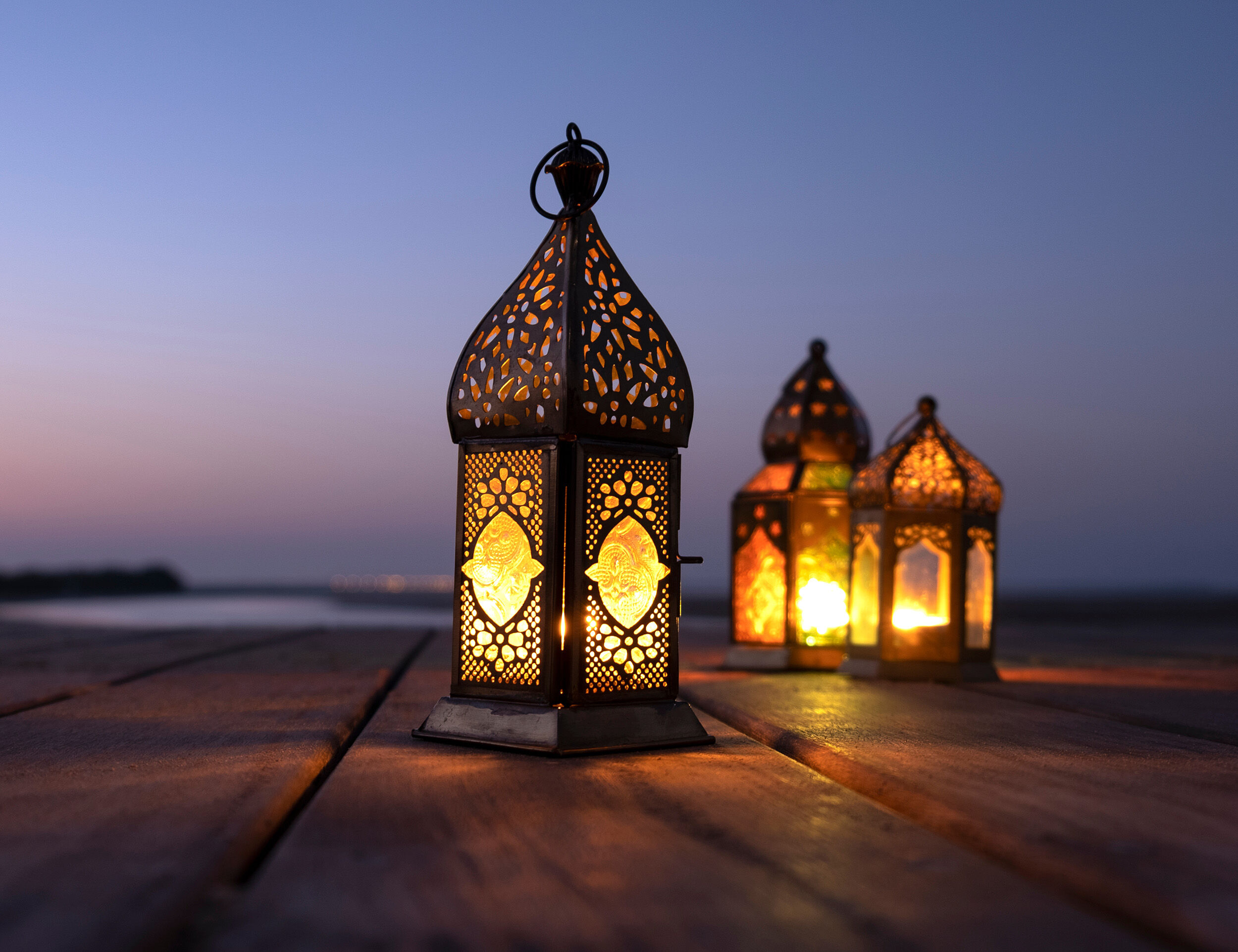 Three glowing lanterns on a wooden pier with a calm sea and twilight sky in the background.
